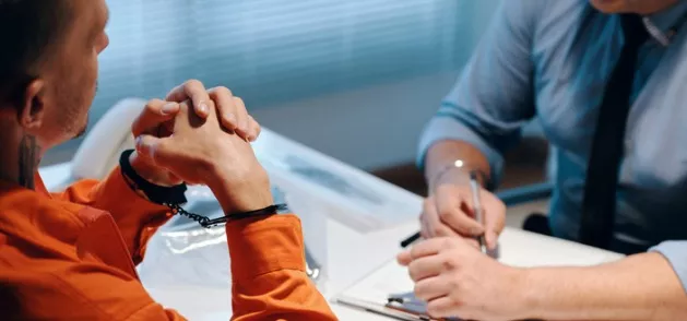 A man in handcuffs talks to a criminal defense lawyer, who is taking notes